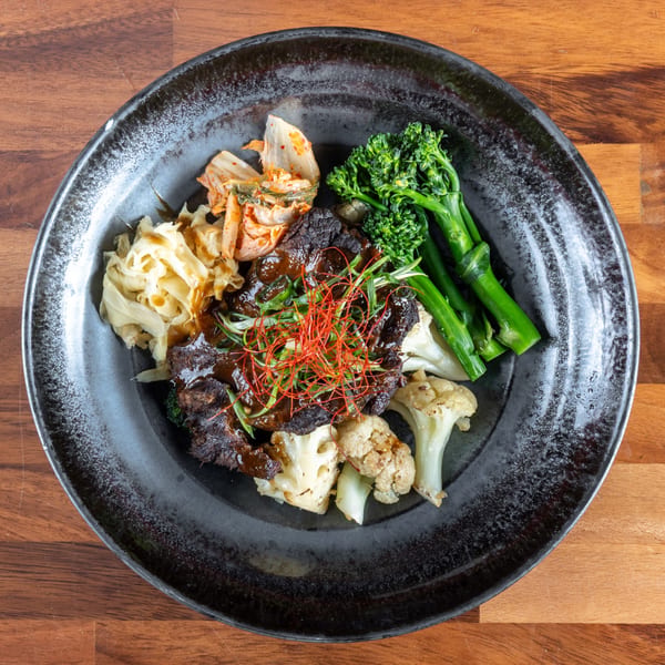 Asian-fusion dish with grilled meat, broccoli, cauliflower, kimchi, and shredded cabbage on a dark plate.