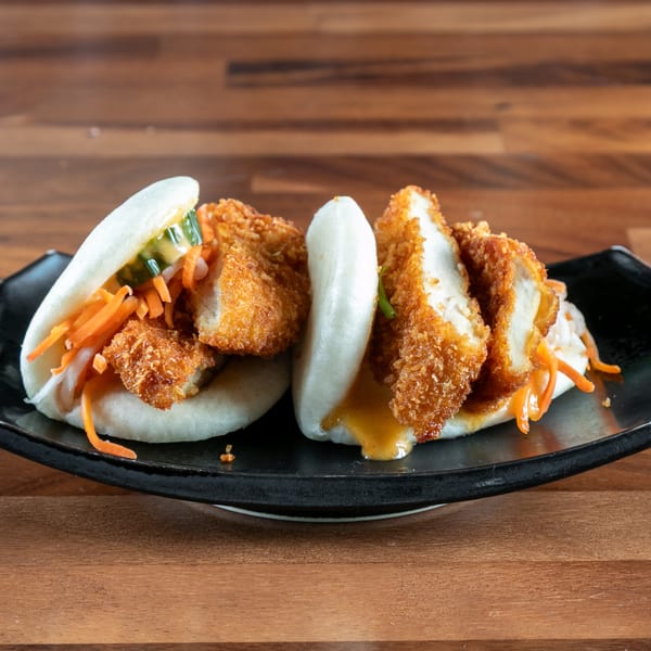 Two Ganzo Sushi bao buns with crispy chicken, pickled vegetables, and sauce on a black plate.