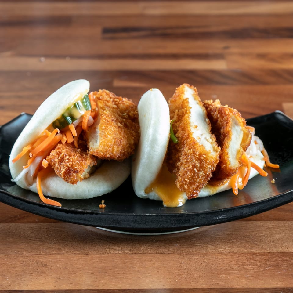 Two Ganzo Sushi bao buns with crispy chicken, pickled vegetables, and sauce on a black plate.
